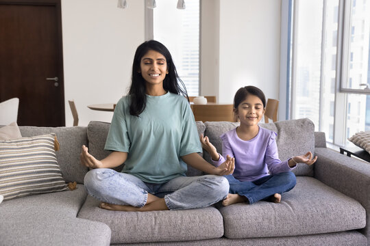 Peaceful Indian mom and preschool daughter meditating with closed eyes at home, doing yoga, sitting on couch, keeping zen fingers, calmness peace hand gestures, smiling