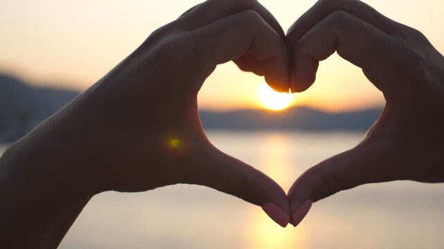Silhouette of young female hands making heart shape over sea background with beautiful sunset. Concept of summer vacation. Close up Slow motion