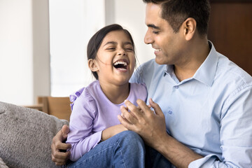 Happy loving Indian dad holding cheerful daughter kid in arms, sitting on home couch, cuddling, tickling laughing girl, having fun with child, enjoying parenthood, family leisure, childcare