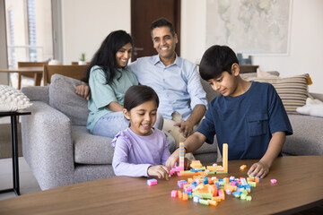 Cute Indian sibling kids playing at home together, constructing toy city towers, developing creative learning skills, stacking buildings. Cheerful parents sitting on couch in blurred background