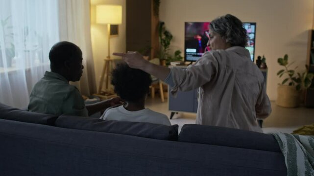 Rear tracking shot of diverse family gathering together in front on TV in evening, dad sitting on sofa and mother with son joining him to watch movie on streaming service in living room