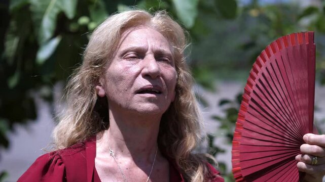 A mature blonde woman fans herself with a handheld fan on a hot sunny day, seeking relief from the heat.