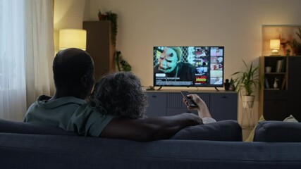 Rear dolly out shot of loving diverse spouses sitting on couch and watching TV after work in evening, husband hugging wife from behind while she choosing movie on streaming service - Powered by Adobe