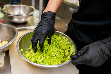 Close-up of hands in black gloves preparing diced vegetables in a kitchen setting, emphasizing...