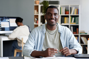 Portrait of smiling African American man holding pen in modern office setting with colleague working in background. Man looking straight at camera while sitting at desk