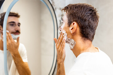 Man with a mustache applies shaving foam in the bathroom in front of the mirror.