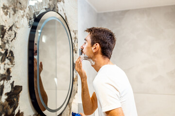 Man with a mustache applies shaving foam in the bathroom in front of the mirror.