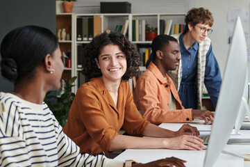 Group of professionals collaborating and discussing work in contemporary office space, with bookshelves and computers visible and diverse individuals working together in casual attire