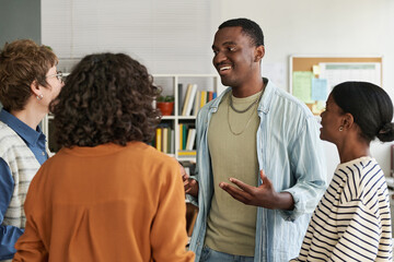 Group of multiracial colleagues laughing during light-hearted conversation in modern office setting with bookshelves and documents in background