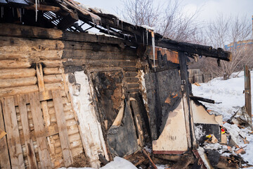 A wooden building shows severe fire damage, with charred walls and a collapsing roof, surrounded by snow. Icicles hang from the remnants, a cold winter day