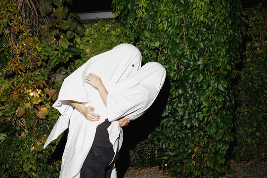 Young couple wearing ghost costumes and kissing each other near plants at Halloween party