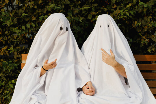 Young couple wearing ghost costumes and talking to each other on bench near plants