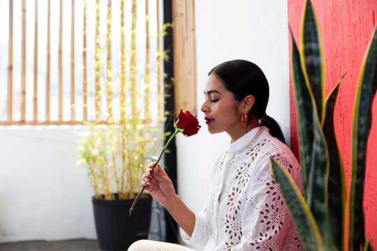 Woman smelling rose flower near plants in front of wall