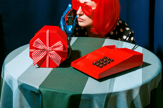 Woman with red wig talking on old-fashioned telephone by gift box on table in studio