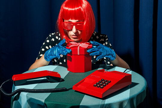 Retro woman with red wig and eyeglasses opening gift box on table in studio
