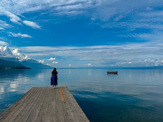 Fototapeta premium Tourist woman modeling on wooden platform with panoramic morning views of Lake Ohrid, North Macedonia. Calm water reflecting small floating boat and surrounding hills and mountains. Tranquil sunrise