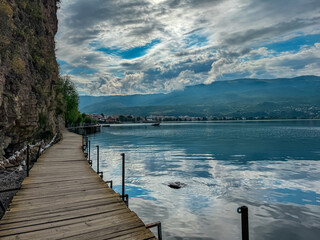 Fototapeta premium Morning walk on wooden platform The Bridge of Wishes extending into calm waters of Lake Ohrid, North Macedonia. Pier boardwalk offers scenic views of tourist lakeside town, rolling hills and mountains