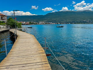 Obraz premium Walking on wooden platform The Bridge of Wishes extending into calm waters of Lake Ohrid in North Macedonia. Pier boardwalk offering scenic views of tourist lakeside town, rolling hills and mountains