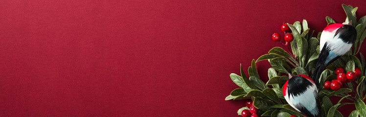 Festive border with bullfinch birds, vibrant red berries, with green foliage on a rich red...