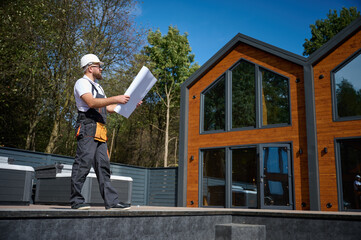 Architect hold plan. Bearded architect wearing hardhat holding project blueprints paper plans standing near comlex of new modern houses looking to something at construction site.