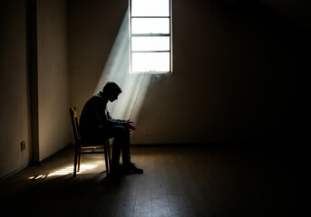 A solitary figure sits in a dimly lit room illuminated by sunlight streaming through a window during the late afternoon