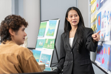 Young businesswoman leading a corporate presentation with global data charts and graphs, explaining business trends and strategies to colleagues during a team meeting in a modern office environment
