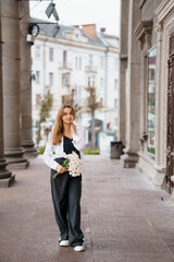 Fototapeta premium Elegant Young Woman in Stylish Attire Holding Flowers in Urban Setting