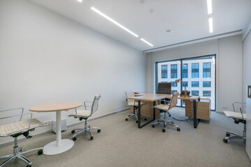 A spacious office features a large window allowing natural light, minimal furniture with a wooden desk, and ergonomic chairs arranged for collaboration.