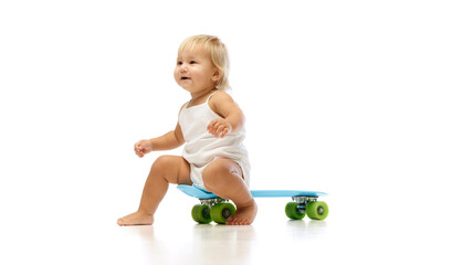 Fototapeta premium Cheerful and beautiful girl, toddler in white onesie sitting on skateboard, playing, isolated on white studio background. Concept of childhood, motherhood, care