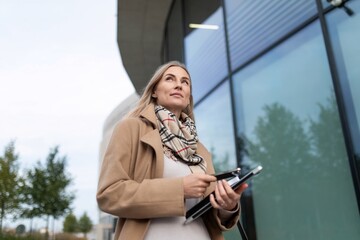 A businesswoman stands outside a business center, holding a laptop and looking up at the sky