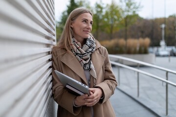 Fototapeta premium A businesswoman stands outside a business center, holding a tablet and looking thoughtfully into the distance