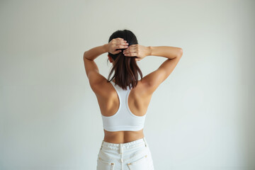 Obraz premium Asian woman with long hair wearing a white tank top and matching pants raising hand arranging hair posing in a bright room with a window