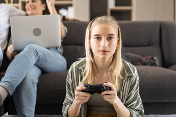 Caucasian girl sitting on the floor controlling joystick playing video game in the living room while parent working on couch. leisure activity on weekend