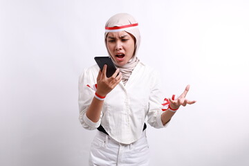 Angry young Asian Muslim woman wearing a hijab shouting loudly at mobile phone isolated over white background. Celebrate Indonesian independence day on 17 August