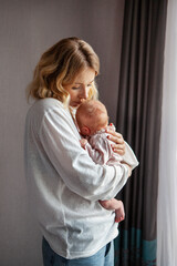 Beautiful blonde mother holds newborn baby in arms dressed in white, standing against minimalist grey wall background. Natural light indoor setting, bond and love between mom and child