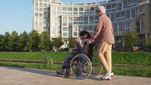 Senior woman in wheelchair walking with caregiver old man on road. Family couple man husband supporting taking care of paralyzed woman wife in chair for people with disability outdoor. Rehabilitation