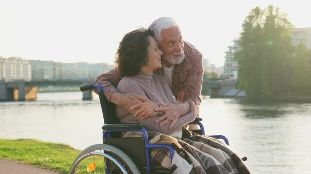 Senior woman in wheelchair walking with caregiver old man on road in park. Elderly family couple man supporting embracing paralyzed woman in chair for people with disability outdoor. Rehabilitation
