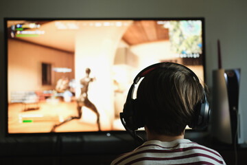 Unrecognizable young boy facing away from the camera. wearing headphones, sitting in front of a large screen playing a video game.