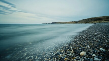 Fototapeta premium A serene seascape with a coastline of smooth pebbles and gentle waves lapping on the shore under a cloudy sky.