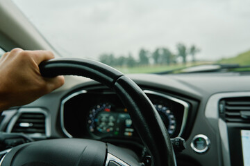 Driver hand gripping steering wheel with view of countryside road ahead through windshield. Man driving car in rainy day. Inside view of modern vehicle interior