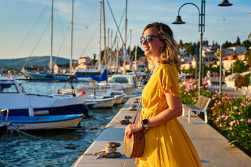 Woman is walking alone on sunny promenade with yacht in sea. Female tourist strolls along marina at...
