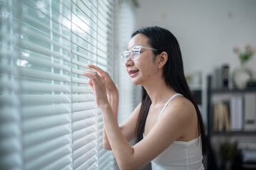 Fototapeta premium Young woman wearing glasses opening blinds and looking out the window