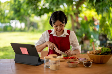 Asian woman following recipe on tablet and cutting vegetables for healthy salad in a green garden