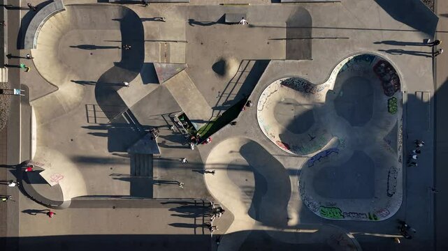 Aerial View of Vilnius Skatepark Next to White Bridge with Skaters and Shadows. Top-down view of a skatepark in Vilnius, Lithuania, showing skaters, ramps, and graffiti-covered bowls.