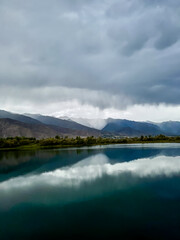 Calm lake waters reflect dark clouds and mountains in the background. The grey sky hangs over the lake, creating a pre-storm atmosphere.
