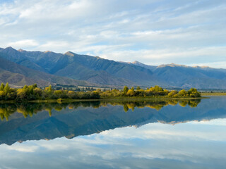 Obraz premium The calm water of a mountain lake reflects the blue sky and white clouds. The green shoreline frames the lake, creating a sense of harmony and tranquility.