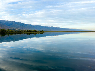 Obraz premium The calm water of a mountain lake reflects the blue sky and white clouds. The green shoreline frames the lake, creating a sense of harmony and tranquility.