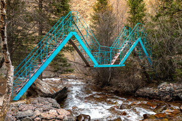 A blue metal bridge with a zigzag design crossing a rocky stream surrounded by trees in a forest....