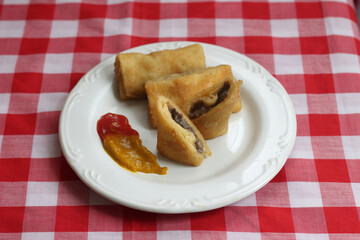 Three rolls of rissoles on a white plate placed on a red-and-white checkered tablecloth. Dutch influenced Indonesian snack. This one with meat and mushroom filling.