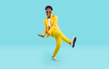 Fototapeta premium Party time. Cheerful, positive and energetic african american woman with funny crazy expression dancing on light blue background. Full length young woman in yellow suit having fun. Fun concept.
